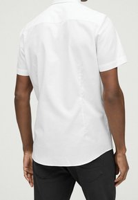 Witte kortarmige shirt met een button-down kraag, gladde katoenstof, getailleerde pasvorm en iets afgeronde zoom. Zwarte broek zichtbaar aan de onderkant.