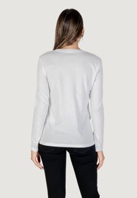 T-shirt bianca a maniche lunghe con scollatura rotonda, tessuto morbido e orlo leggermente curvo, indossata con pantaloni neri.
