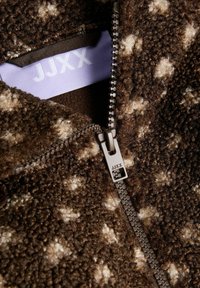 Primo piano di una giacca in pile marrone con pois crema, una zip metallica con cursore marchiato e un tessuto morbido e strutturato.