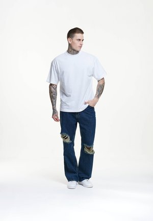 Weißes Baumwoll-T-Shirt mit kurzen Ärmeln, kombiniert mit blauen Jeans, die an den Knien beschädigte Stellen aufweisen. Weiße Sneakers runden den Look ab.