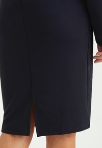 Marineblaues knielanges Kleid aus dehnbarem Stoff, mit einem Schlitz hinten für Bewegungsfreiheit und einer glatten Textur. Einfaches Design ohne Muster.