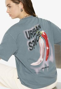 Graues Baumwoll-T-Shirt mit übergroßem Grafikdruck von roten und weißen Chilischoten, "URBAN SPICE" Text und "HARLEM SOUL" Etikett am unteren Ende.