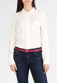 Hvit skjorte med lange ermer, knappelukking og lomme, kombinert med mørk denimbukser og et lyst rosa belte med en klassisk spenne.