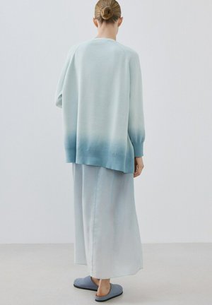 Osoba s kosom u pundžu nosi svijetloplavi ombre džemper, dugu blijedoplavu suknju i plave papuče, stojeći ispred jednobojne pozadine.