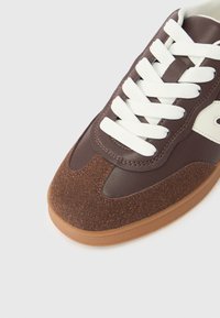 Καφέ αθλητικό παπούτσι με υφασμένη καφέ μύτη, λευκά κορδόνια και απαλό δερμάτινο ε πάνω μέρος. Διαθέτει σόλα από καουτσούκ τύπου γκαμ και ομαλό σχεδιασμό.