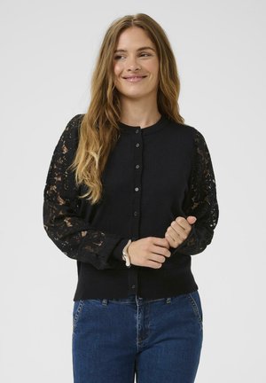 Black cardigan with textured lace long sleeves, round neckline, and button closure. Paired with blue denim. Smooth knit body.