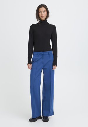 Frau steht und trägt einen schwarzen Rollkragenpullover, weite blaue Hosen und schwarze Schuhe vor einem schlichten hellgrauen Hintergrund.