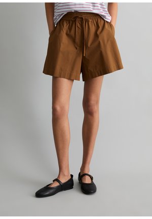 Personne portant un short marron à cordon, un haut rayé partiellement visible et des ballerines Mary Jane noires, debout devant un fond gris uni.