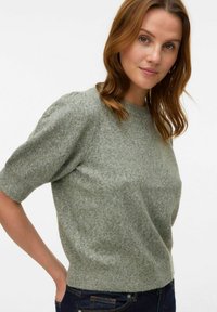 Maglione a maglia verde chiaro con maniche a sbuffo corte, colletto rotondo, texture morbida, vita aderente, abbinato a jeans in denim scuro.