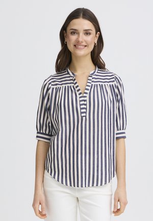 Frau mit schulterlangem braunem Haar, die eine marineblaue und weiße Bluse mit vertikalen Streifen und Ellbogenlangen Ärmeln sowie weiße Hosen trägt, lächelnd.