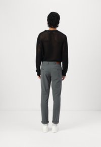 Uomo in piedi con la schiena rivolta, che indossa un maglione in rete nero, pantaloni grigi arrotolati e sneakers bianche, su uno sfondo bianco semplice.