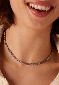 Close-up van een glimlachende vrouw met een zilveren gedraaide ketting om haar hals, kort bruin haar en een beige textuurtop.