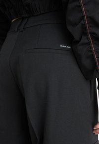 Čierne nohavice s textúrovaným povrchom, ktoré majú zadné vrecko a malú štítok s logom "Calvin Klein". Páska má elastický dizajn.
