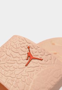 Κοντινό πλάνο λουριού σανδάλου σε μπεζ χρώμα με υφή, που διαθέτει το πορτοκαλί λογότυπο Jumpman εγχάρακτο στην επιφάνεια.