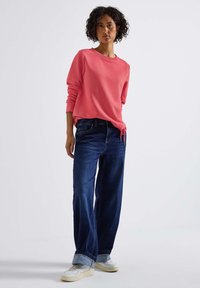 Koraalkleurig shirt met lange mouwen en een ronde hals, gecombineerd met wijdvallende donkerblauwe jeans met een opgestikte zoom en witte sneakers.