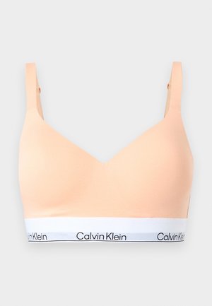 Sutien fără cusături de culoare piersică deschisă, cu bretele reglabile și o bandă elastică albă cu logo-ul „Calvin Klein” în litere negre.