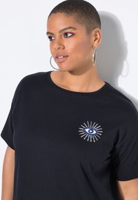Persoon met kort haar en ringoorbellen, gekleed in een zwart t-shirt met een blauw oog en zilveren stralen ontwerp op de borst, kijkt opzij.