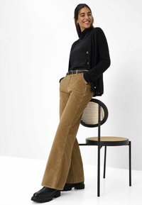 Pantaloni a zampa d'elefante in velluto a coste marrone abbinati a un dolcevita nero e un cardigan, su uno sfondo bianco con una sedia nera.