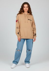 Felpa beige con cappuccio con testo "Amour", grafiche sulle maniche, tasca a marsupio, abbinata a jeans strappati azzurri chiari e sneakers bianche.