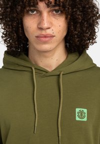 Olive green hoodie featuring a drawstring hood, textured fabric, and a small light green logo on the left chest.