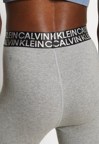 Grå leggings med en glat tekstur, udstyret med en bred sort elastik i taljen præget med "CALVIN KLEIN" i metallisk skrift.