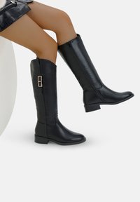 Botas negras hasta la rodilla hechas de cuero suave, con un cierre lateral y una hebilla decorativa en la parte trasera. Tacón bajo en bloque.