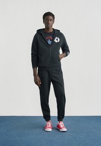 Μαύρο φούτερ με φερμουάρ και μπροστινές τσέπες, φορεμένο πάνω από ένα κόκκινο T-shirt με γραφικό σχέδιο, συνδυασμένο με μαύρο παντελόνι jogger και κόκκινα αθλητικά παπούτσια χαμηλού ύψους.