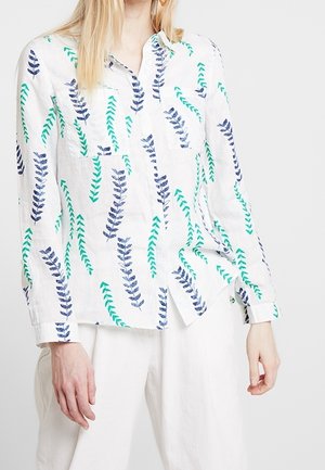 Kobieta ubrana w białą koszulę na guziki z długim rękawem z niebiesko-zielonym wzorem liści, w połączeniu z białymi spodniami.