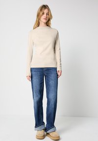 Beżowa bluzka z długim rękawem zestawiona z niebieskimi dżinsami i brązowymi butami z futerkiem. Dżinsy mają podwinięte mankiety, co tworzy luźny krój.