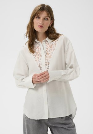 Bela srajca z čipkasto vložkom po sredini. Ima ovratnik z gumbi, rebrasto teksturo in dolge rokave. Kombinirana s sivimi hlačami.