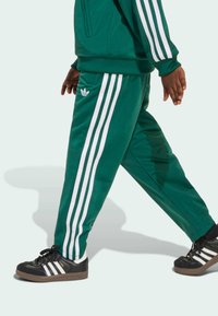 Zelene atletske trenirke s tri bijele vertikalne pruge, s malim logotipom na džepu, uparenim s crnim tenisicama s bijelim prugama.