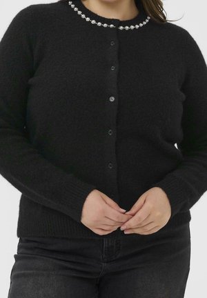 Kvinde iført sort knaplukket fluffy cardigan med juvelbesat udskæring, hænder foldet foran, mod en ensfarvet lys baggrund.