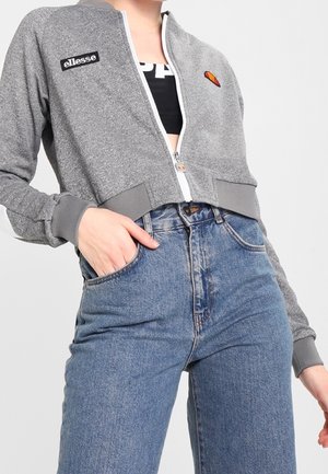 Personne portant une veste courte grise zippée avec le logo ellesse et un jean bleu taille haute, main dans la poche avant.