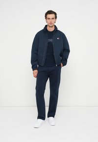 Giacca bomber blu sopra una maglietta blu con logo, abbinata a pantaloni blu e sneakers bianche; tessuto liscio, vestibilità casual.