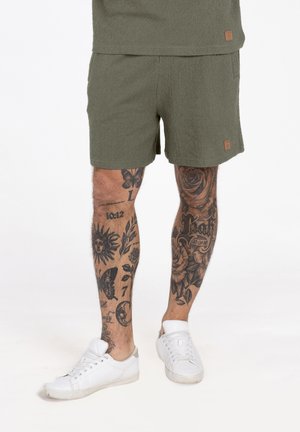 Osoba ubrana w oliwkowozielone szorty i białe tenisówki, z szczegółowymi tatuażami z czarnego tuszu pokrywającymi oba dolne nogi.