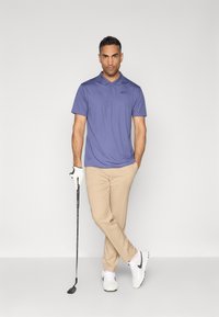 Polo tričko vo svetlo fialovej farbe, krátke rukávy, s čiernym logom. Béžové nohavice. Biele golfové topánky, drží golfovú palicu, s bielou rukavicou.