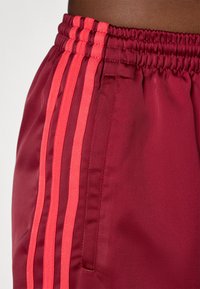 Bordo sportiniai šortai su lygiu paviršiumi, su trimis vertikaliomis rožinėmis juostelėmis šonuose ir elastinga juosmens juosta.
