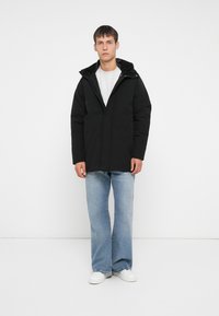 Svart jakke med hette, med glidelås foran og to lommer foran. Kombinert med lyseblå utsvingte jeans og hvite sneakers.