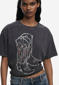 Un t-shirt gris surdimensionné avec un graphic détaillé de botte de cowboy, rehaussé de touches rouges et blanches. Manches courtes et encolure décontractée, associé à un jean en denim.