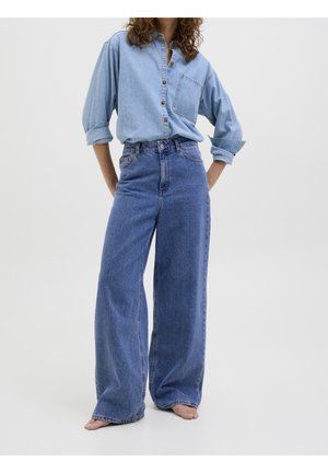 Personne portant une chemise en jean bleu clair aux manches retroussées, rentrée dans un jean bleu taille haute à jambes larges, debout pieds nus sur un sol blanc.