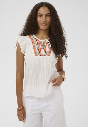 Mujer con cabello largo y rizado, vistiendo una blusa blanca sin mangas con bordados en naranja y verde, y pantalones blancos, de pie con una mano en el bolsillo.