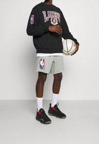 Sudadera negra con "LAKERS" en purpura, pantalones cortos atléticos grises con el logo de la NBA, zapatillas negras con acentos rojos, sosteniendo un balón de baloncesto.