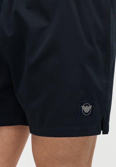 Κοντινή λήψη μαύρων υφασμάτινων σορτς στον μηρό ενός ατόμου με ένα μικρό ορθογώνιο έμβλημα κοντά στο τελείωμα.