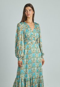 Robe longue turquoise avec une silhouette fluide. Elle présente un décolleté en V, de longues manches bouffantes, un tissu à motifs bleu, orange et blanc, et une taille ceinturée.