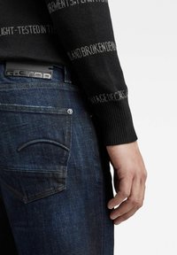 Suéter negro con patrón texturizado y letras grises, combinado con jeans de mezclilla azul oscuro que presentan un diseño clásico de bolsillos y etiqueta de marca.