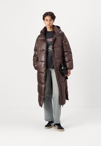 Abrigo puffer largo marrón con acabado brillante, que incluye capucha, bolsillos laterales y cierre de cremallera; llevado sobre una camiseta gráfica negra y jeans.