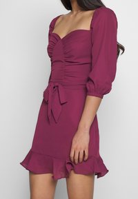 Kortärmad, figurnära klänning i djup plommon med rynkad byst, knytband vid midjan och ryschdetaljer i fållen. Slät tygtextur.