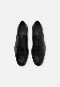 Zapatos de cuero negro con acabado suave, punta redondeada, diseño de cordones y un detalle de puntera en contraste. Suela plana, diseño minimalista.