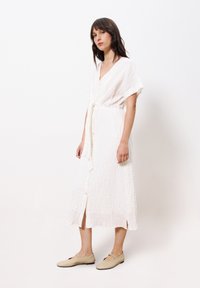 Weißes, knielanges Kleid aus strukturiertem Stoff, mit V-Ausschnitt, kurzen Ärmeln und gebundenem Taillengürtel; hat Knopfdetails und seitliche Schlitze.