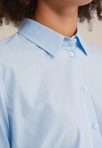 Primo piano di una persona che indossa una camicia azzurro chiaro con colletto a punta e quattro bottoni bianchi ben visibili.